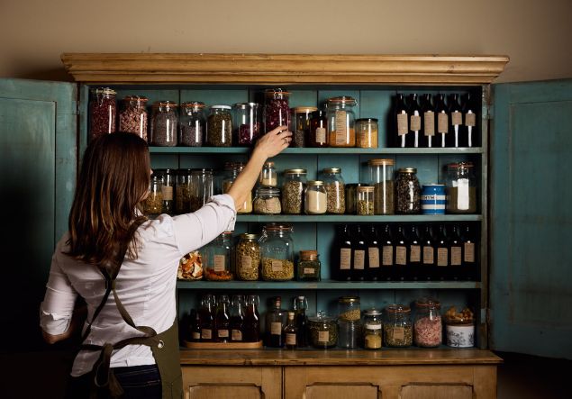 A woman reaches for a herbal remedy in front of an open cupboard full of glass jars full of herbs