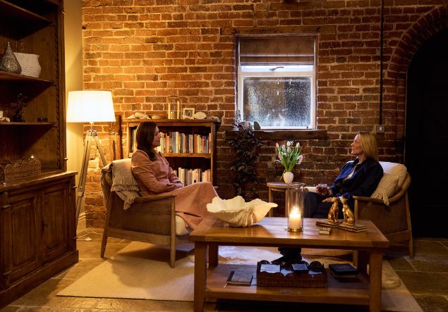Womens wellness 1 A woman in a pink dress is explaining something from a document in her hand to another woman. They are in a warm room with candles and wooden furniture